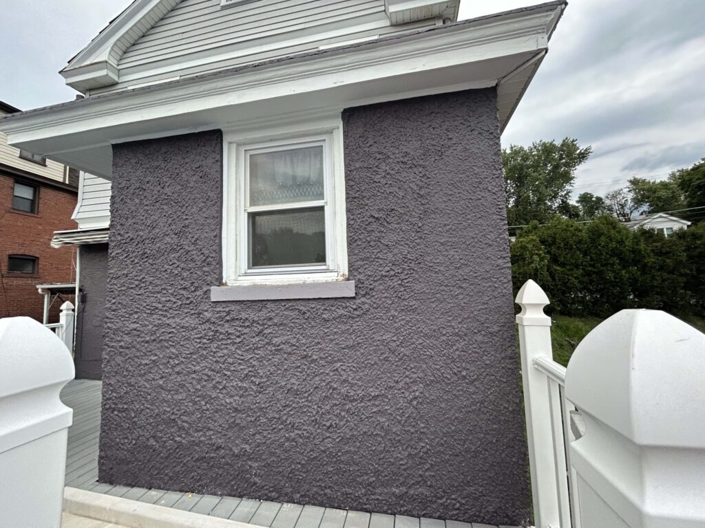 A house exterior with a textured gray wall, a single window, and white trim, next to a white fence and greenery.