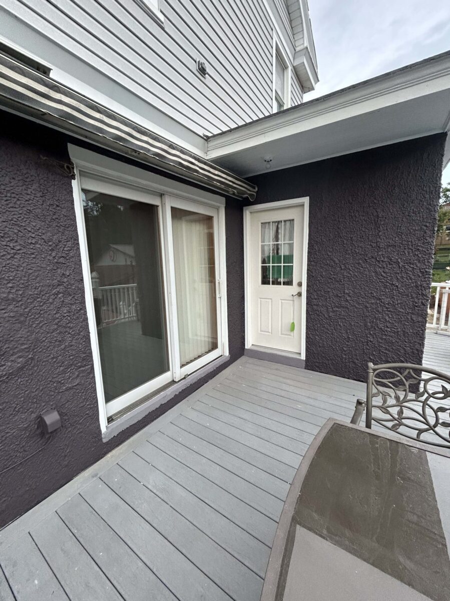 A small outdoor deck with gray flooring, a glass table, sliding doors, and a single white door against a dark wall.